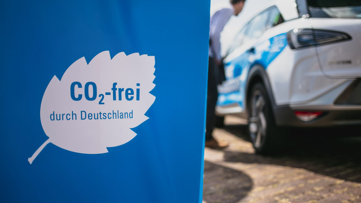 Fahne auf der steht CO2-frei durch Deutschland, im Hintergrund parkt ein Wasserstoff-Pkw